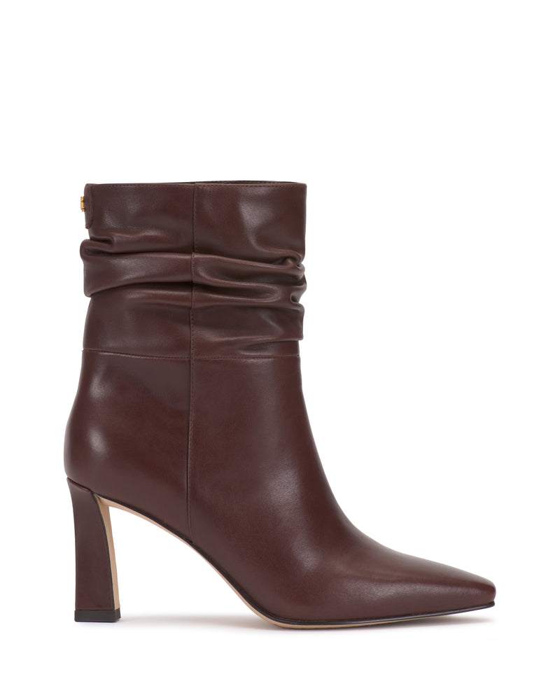 The Vince Camuto Grenna Slouchy Bootie is featured in side view on a white background, showcasing its brown leather construction, pointed toe, mid-height heel, and slouched ankle shaft detail.