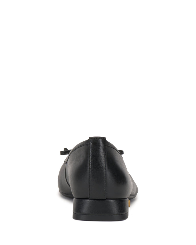 The Vince Camuto Jenlea Ballet Flat is shown from the back on a white background. This black leather shoe features a low block heel, smooth design, and a delicate bow accent on top.