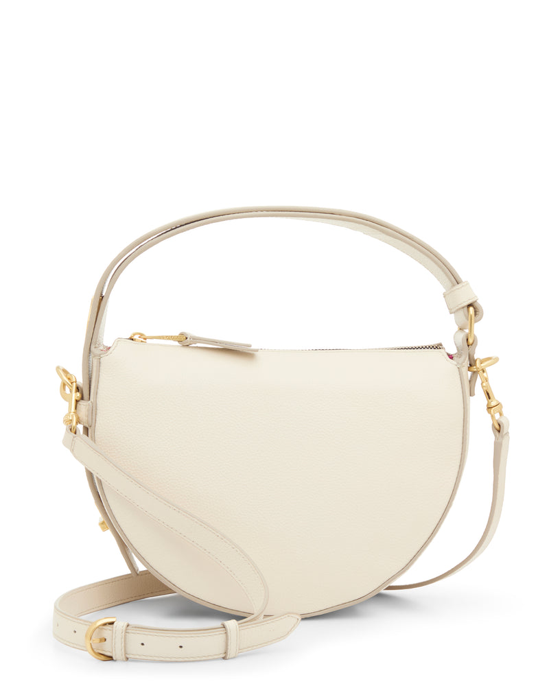 The Vince Camuto Karla Crossbody Bag is cream-colored, half-moon shaped, made of leather with a zip top, gold hardware, and an adjustable strap, shown on a white background.