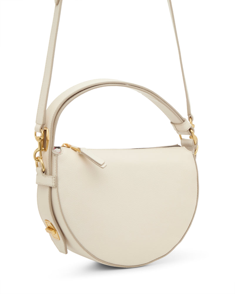 The Vince Camuto Karla Crossbody Bag is a cream, round leather handbag with a top handle and long shoulder strap. It features gold hardware, a zipper closure, and a minimalist design.