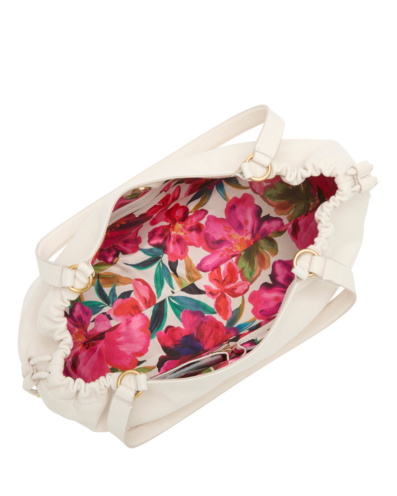 The Leane Tote Bag by Vince Camuto is a white handbag with a drawstring opening, shown from above. Its interior has a vibrant pink, red, and green floral lining, and gold hardware accents decorate the handles.