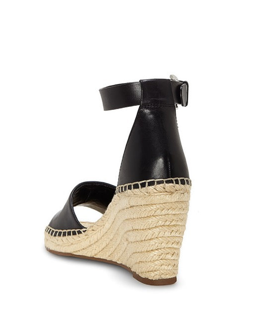 The Vince Camuto Leera Espadrille Wedge Sandal is shown from the back on a white background, featuring a black ankle strap, open toe design, and a woven espadrille wedge heel.