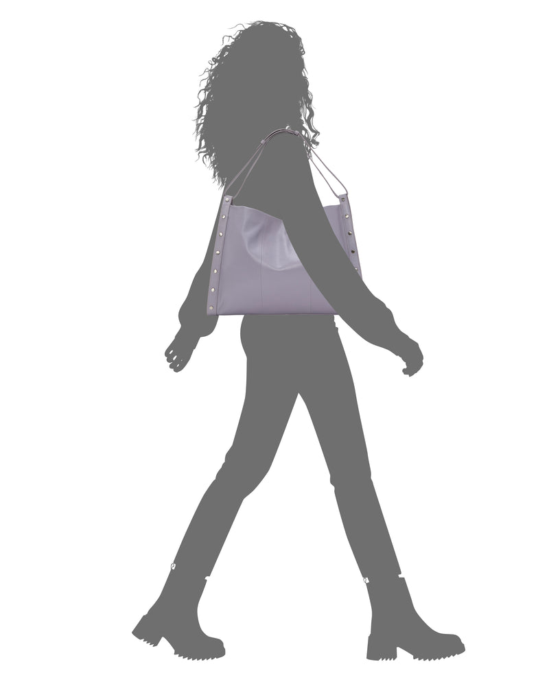 Silhouette of a woman carrying the Vince Camuto Lisha Tote Bag—a spacious, light purple tote with stud details—over her shoulder. She wears boots, pants, and a jacket against a white background.