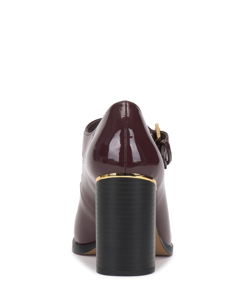A rear view of the Vince Camuto Micha Pump in glossy burgundy showcases its T-strap design, thick black heel, and gold accent near the base—all displayed on a white background. This high-heeled shoe effortlessly combines vintage charm with feminine style.