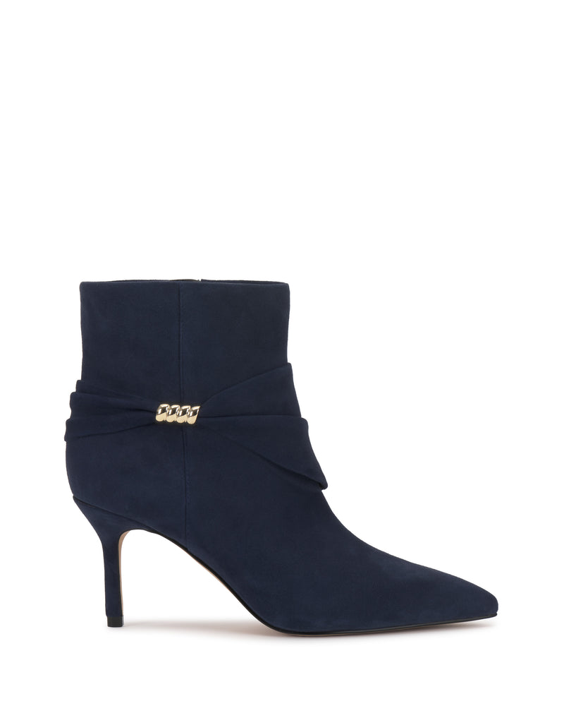 The Murlea Bootie by Vince Camuto is a navy blue suede ankle boot with a pointed toe, mid stiletto heel, and folded wrap detail adorned with a small gold chain accent.