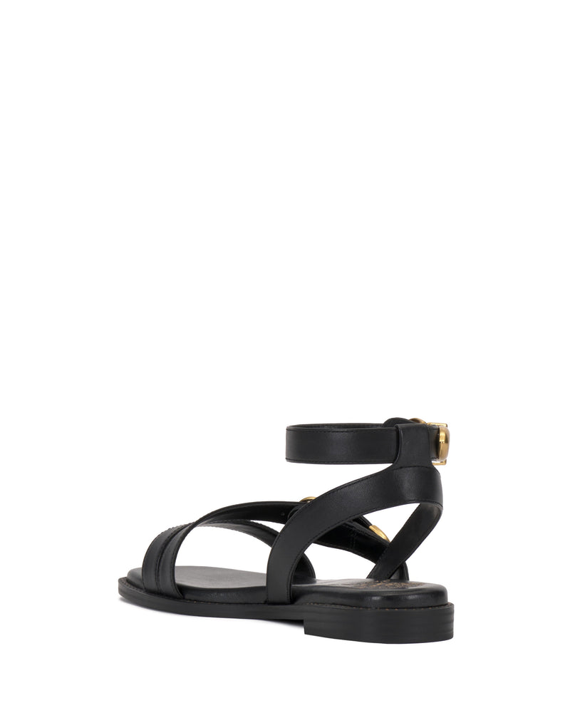 The Vince Camuto Neiko Sandal is a black flat with crisscross straps and an adjustable gold-buckle ankle strap, shown from the back-left side on a white background.