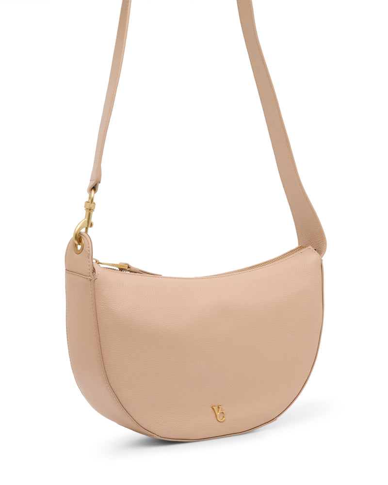 The Vince Camuto Orlie Crossbody Bag, shown against a white background, is crafted from beige leather and features a gold zipper along with a small gold logo on the lower front. This chic accessory effortlessly blends elegance with everyday functionality.