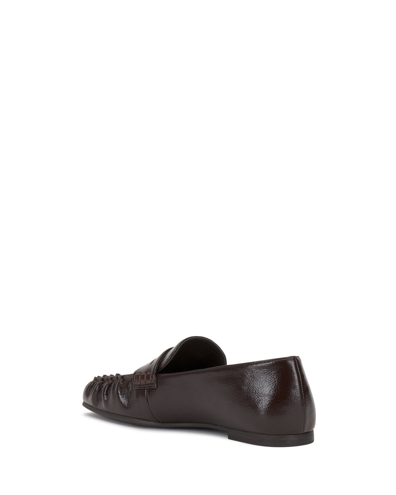 The Vince Camuto Paelyn Penny Loafer is shown from a rear side angle on a white background, showcasing its moccasin-inspired textured front and smooth dark brown leather heel and sides.