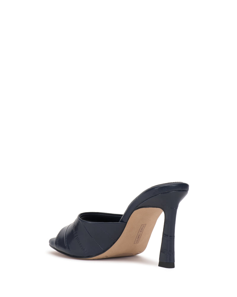 The Vince Camuto Paigely Mule is a black minimalist shoe with an open toe, quilted texture, and beige sole, featuring a flared heel. It is shown from the back at an angle against a white background.