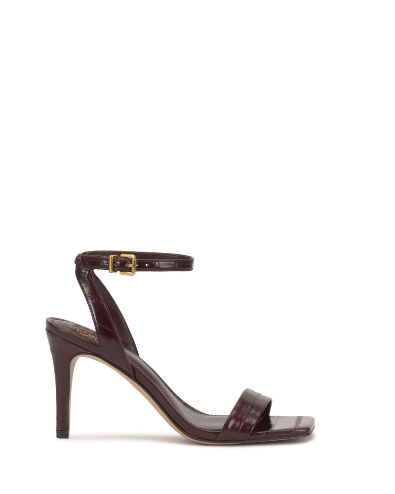 The Vince Camuto Saprenda Sandal is a dark burgundy metallic leather dress sandal featuring an open toe, single front strap, slim high heel, and ankle strap with a gold buckle, shown on a white background.