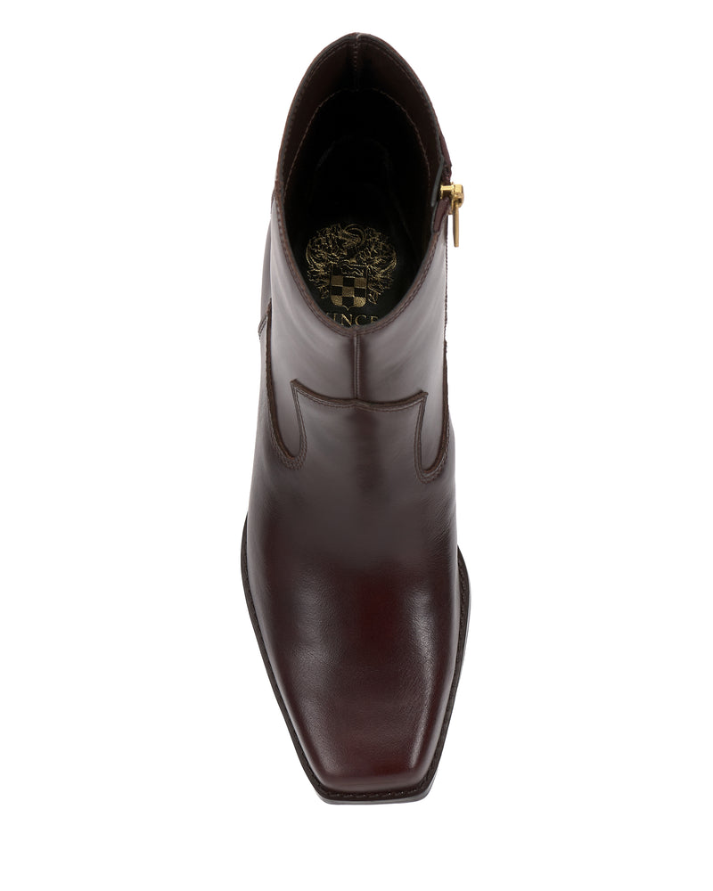 A top view of a single Sybren Bootie by Vince Camuto in dark brown leather features a square toe, side zipper, and a gold emblem with a crown and shield on the insole. This design exudes modern sophistication and is finished with a chunky block heel that offers both style and stability.