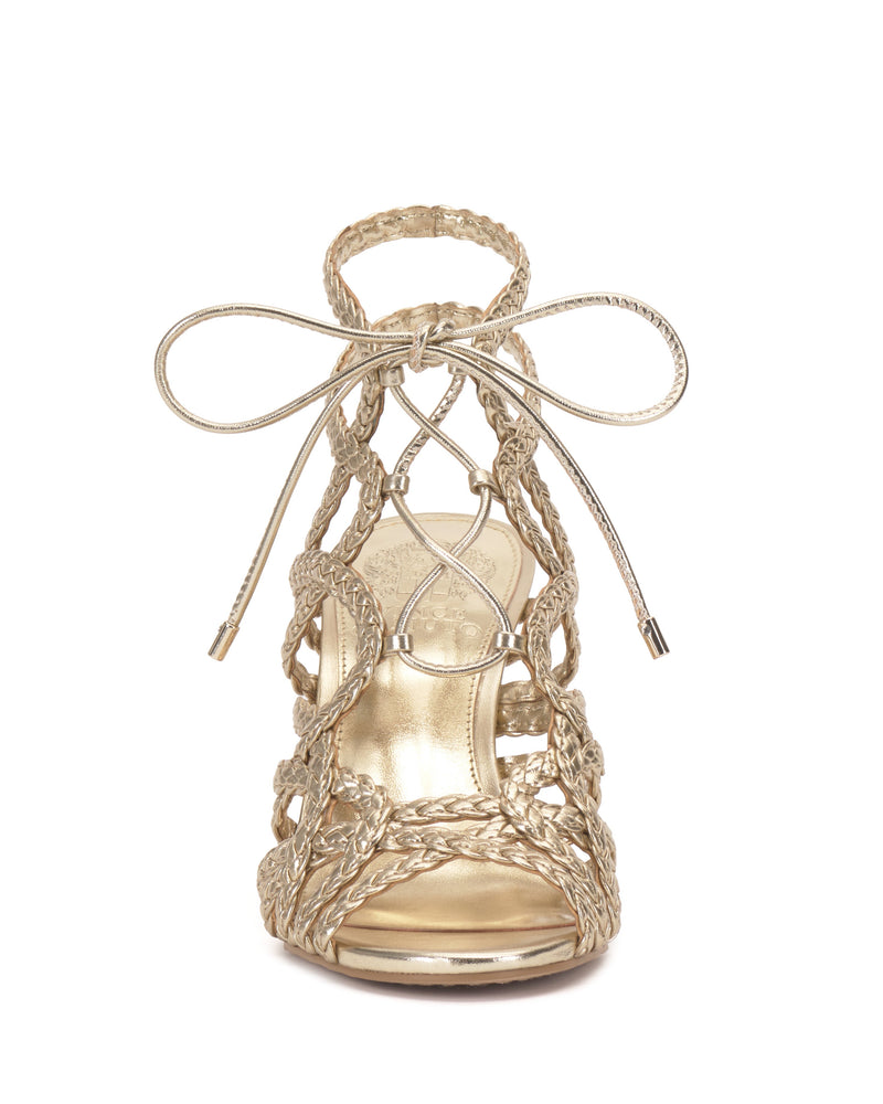 The Tilly Sandal by Vince Camuto features a front view of gold strappy stilettos with intricate braided straps, a decorative ankle bow, open toe design, and a shiny metallic finish.