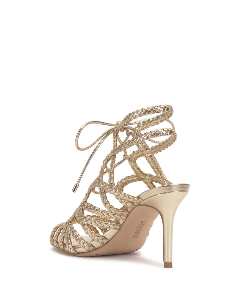 The Vince Camuto Tilly Sandal is a gold strappy sandal with braided straps, an open back, chic lace-up ankle design, and a slim stiletto heel. Displayed at an angle, it highlights both the elegant heel and side profile.