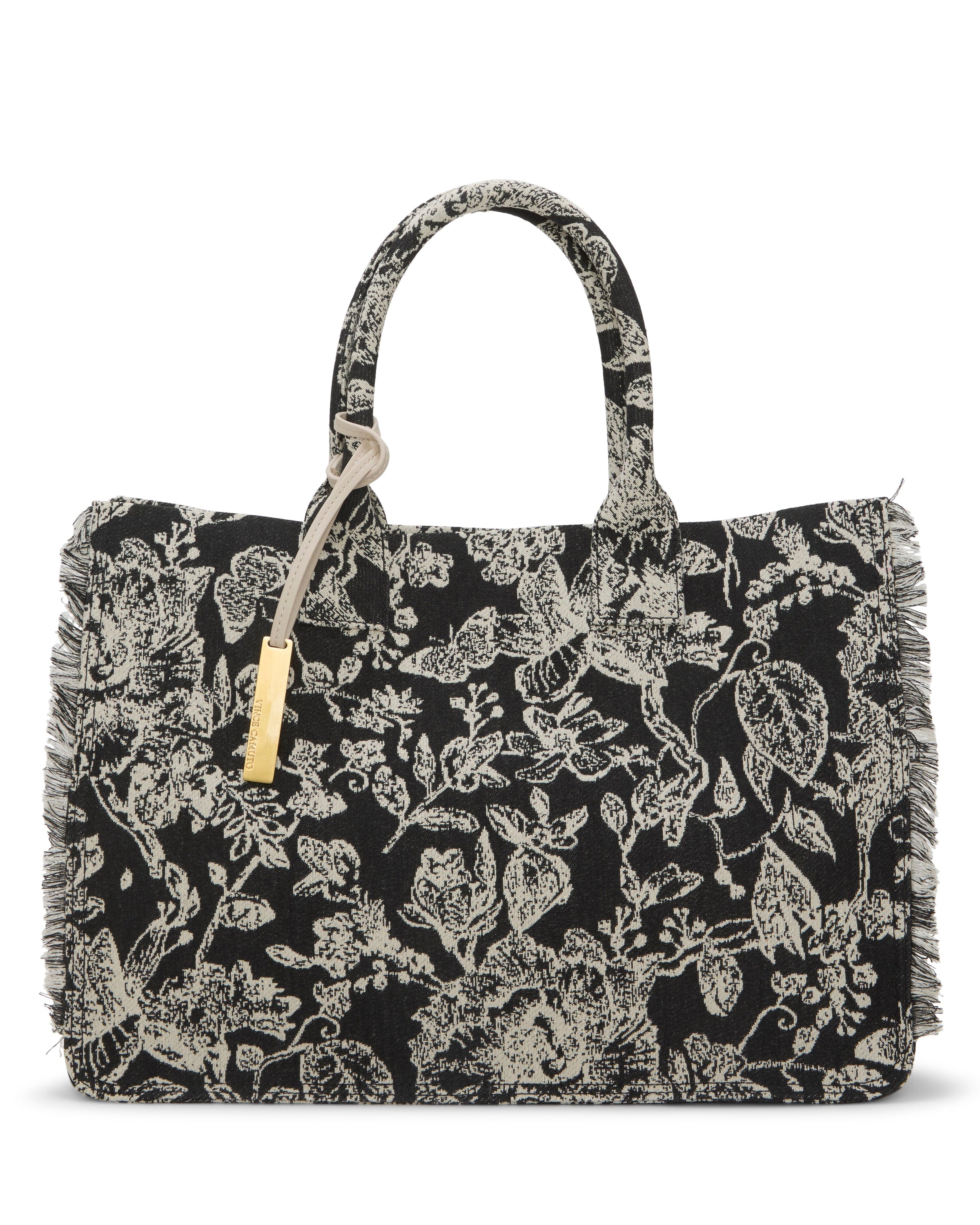 The Vince Camuto Zhury Tote Bag is a black tote with off-white floral and bird patterns, two handles, fringe trim along the edges, and a gold-tone tag hanging from one handle.