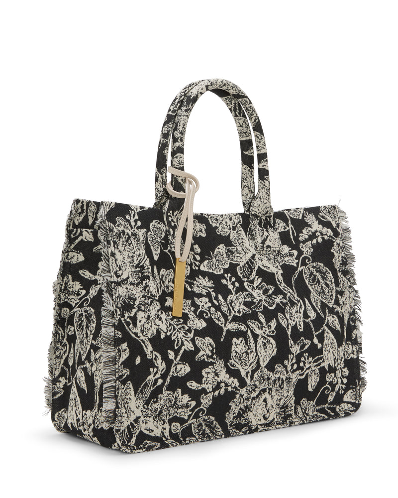 The Vince Camuto Zhury Tote Bag features a black background with white floral patterns, two handles, a rectangular shape, slightly frayed edges, and a beige tag with a gold accent for added charm.