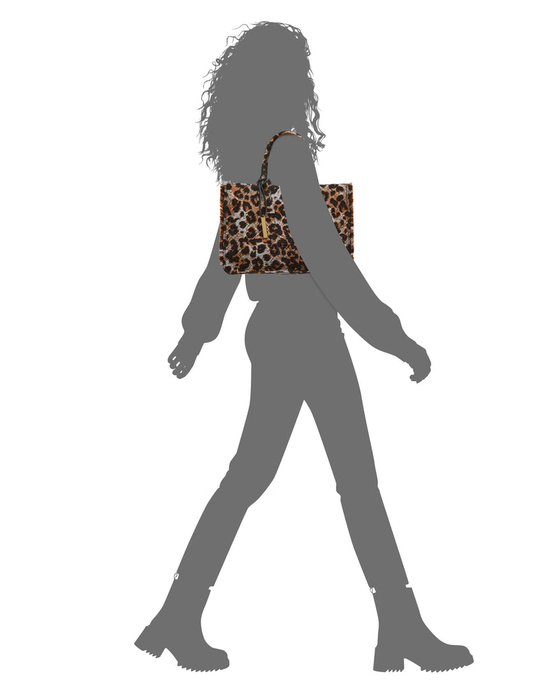 Silhouette of a person walking, carrying the Vince Camuto Zhury Tote Bag with a leopard print on their shoulder. The person has curly hair and is wearing boots and pants.