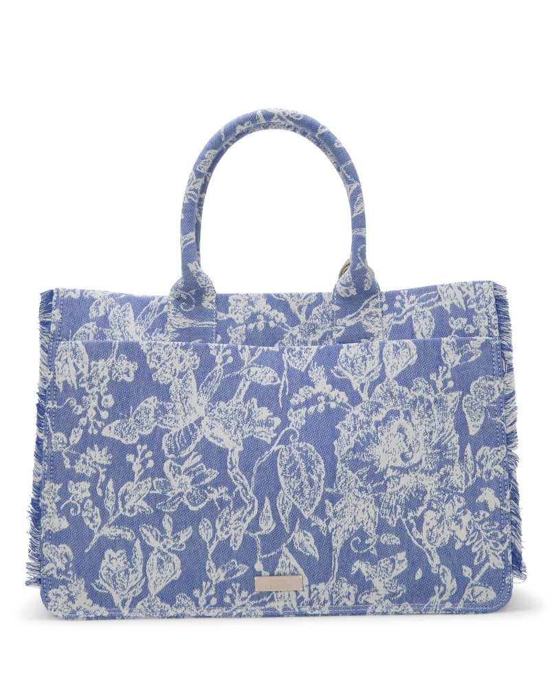 The Vince Camuto Zhury Tote Bag is blue with white floral and animal prints, fringed edges, two handles, and a small metal label on the front near the base.