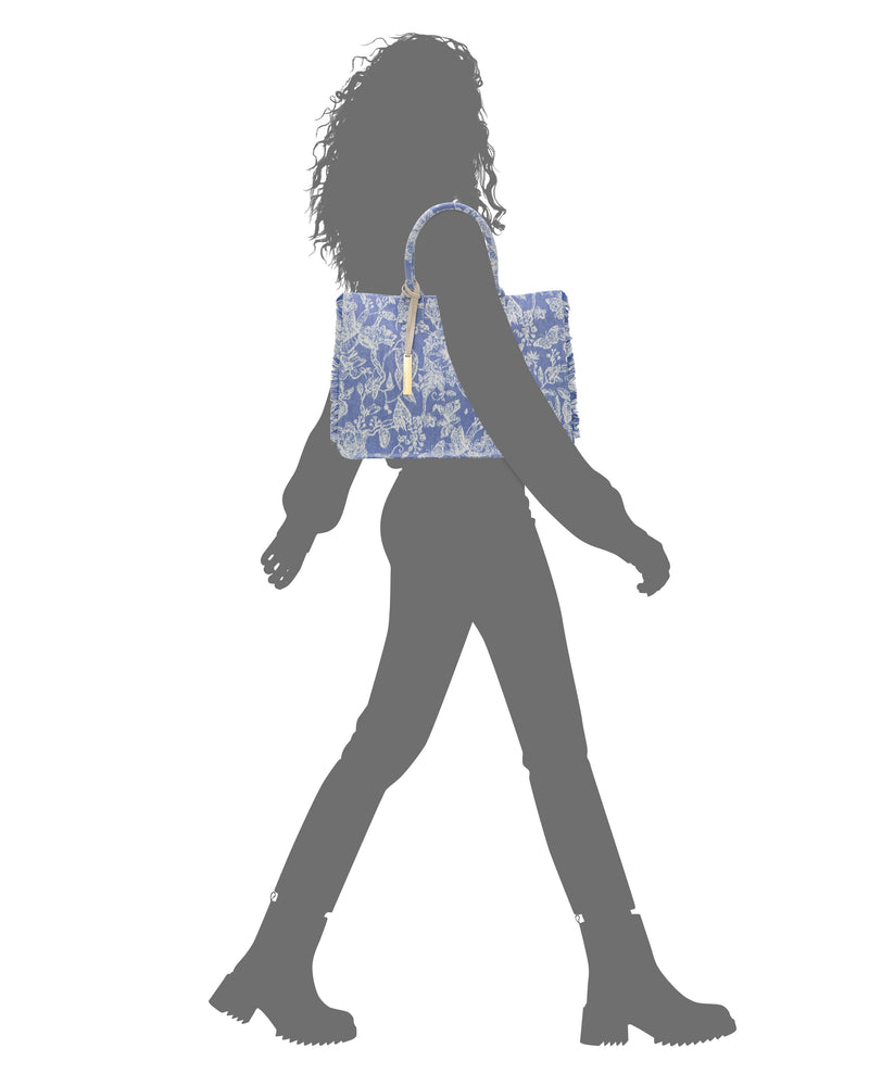 Silhouette of a person with curly hair walking in boots, carrying the Vince Camuto Zhury Tote Bag—large, blue and white patterned—over their shoulder.