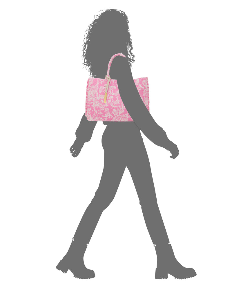 A person with curly hair walks in boots and fitted pants, carrying a pink, patterned Vince Camuto Zhury Tote Bag on their shoulder.