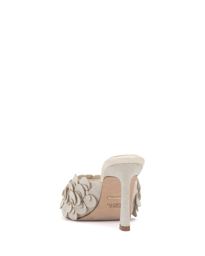 The Rose Mule by Vince Camuto is a beige 3D blossom mule with an open toe, textured fabric, floral appliqué on the strap, and a light sole, shown from the back.