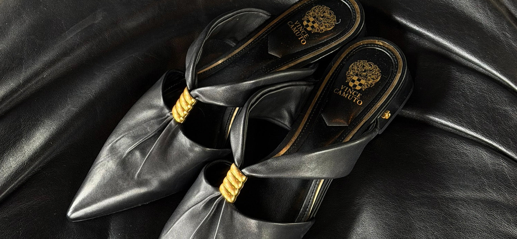 A pair of black leather pointed-toe mule flats with gold trim and a gold Versace logo on the insole, displayed on a black leather background.