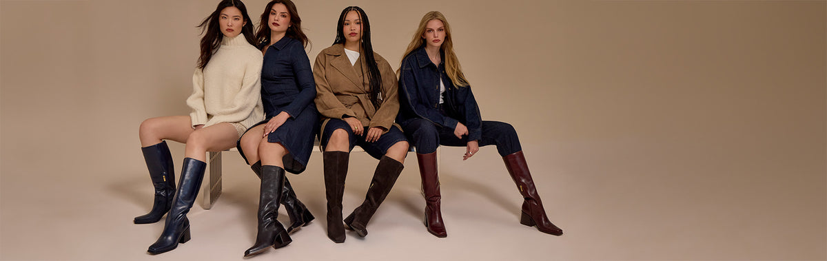 Four women seated side by side wearing long over-the-knee boots in different colors and styles, posed against a plain beige studio background.
