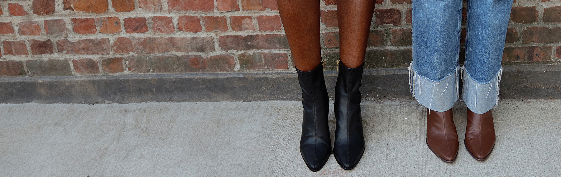 Two models' lower legs standing side by side against a brick wall, showcasing different styles of ankle boots. The model on the left wears black leather pointed-toe ankle boots, while the model on the right wears brown leather ankle boots with cuffed blue jeans.