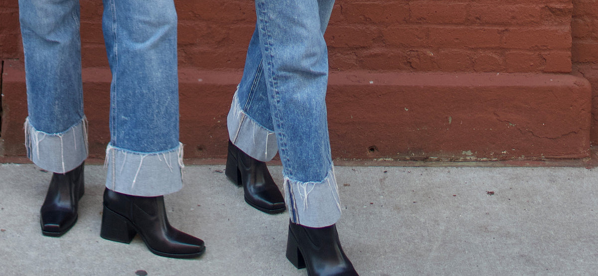 close up of black square toe boots and jeans on a street against a red brick wall 