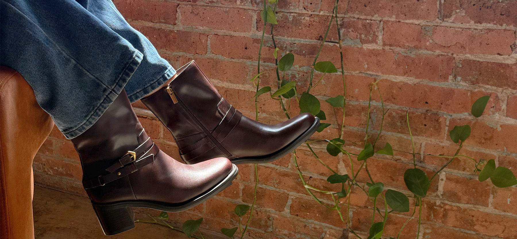 A person wearing dark brown leather ankle boots with blue jeans, sitting on a wooden chair in front of a red brick wall with green trailing vines.