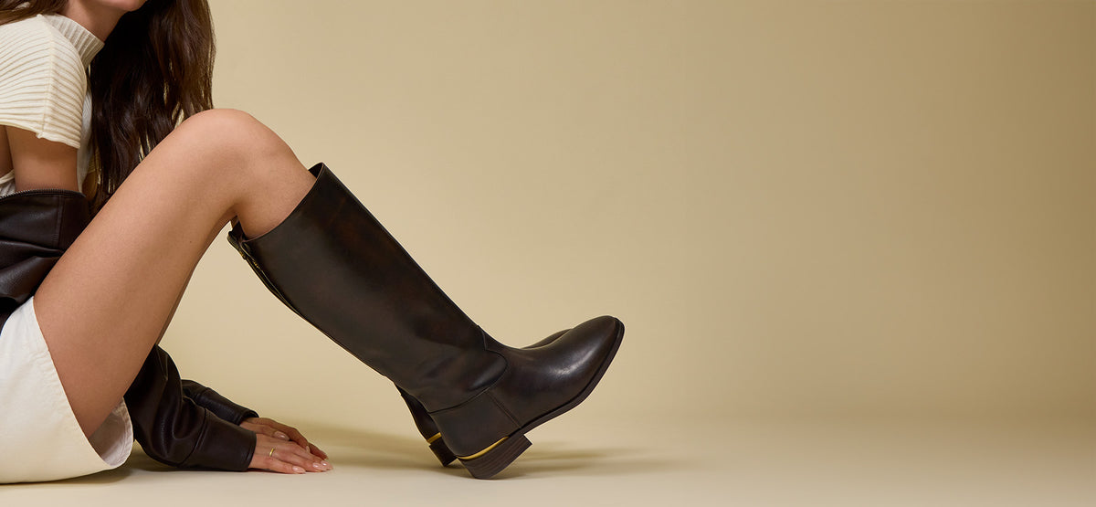 Close-up of woman in dark brown knee-high boots and gold detailing sitting with her knees up against a beige studio background.