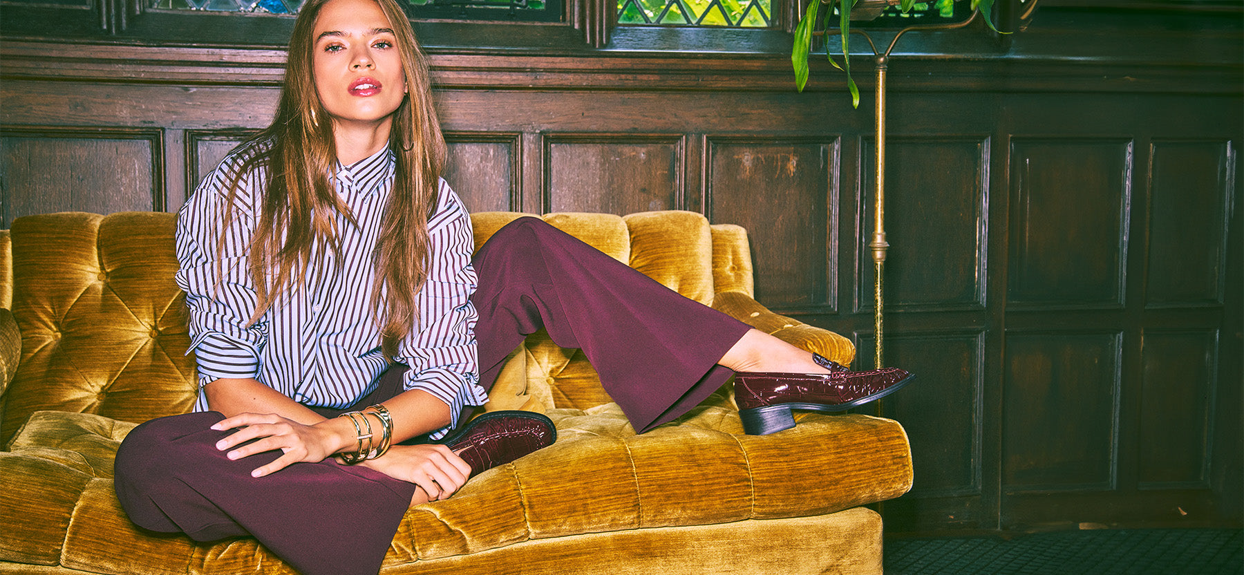 A woman sits on a mustard-yellow velvet sofa in a dark wood-paneled room, wearing a blue and white striped button-up shirt, dark burgundy wide-leg trousers, and dark burgundy heeled shoes.