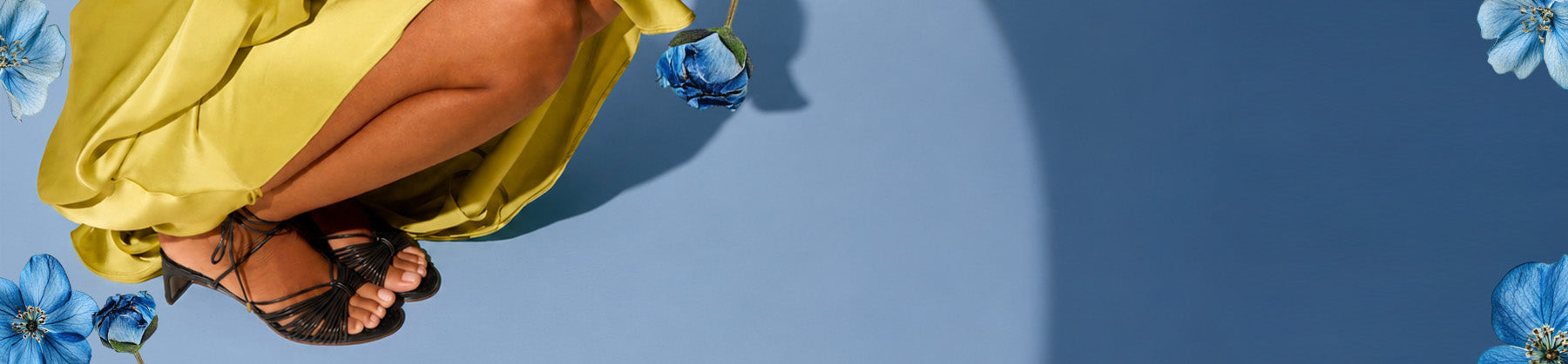 A person wearing a flowing yellow dress and black strappy sandals, seated against a blue background with decorative blue flowers at the corners.