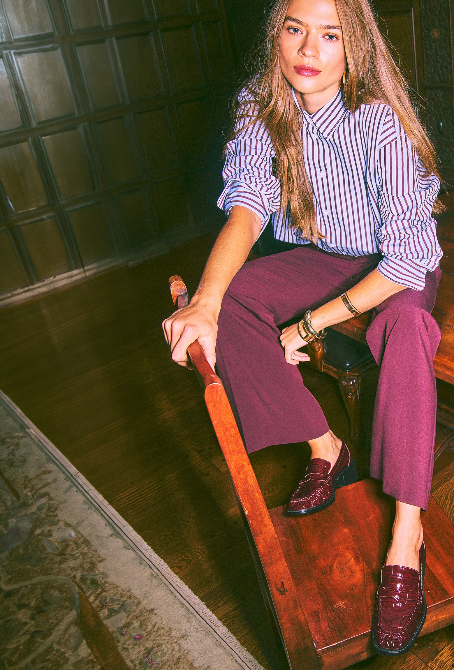 A woman sits on a wooden bench in a dimly lit room, wearing a blue and white vertically striped button-down shirt, burgundy wide-leg trousers, and burgundy loafers.