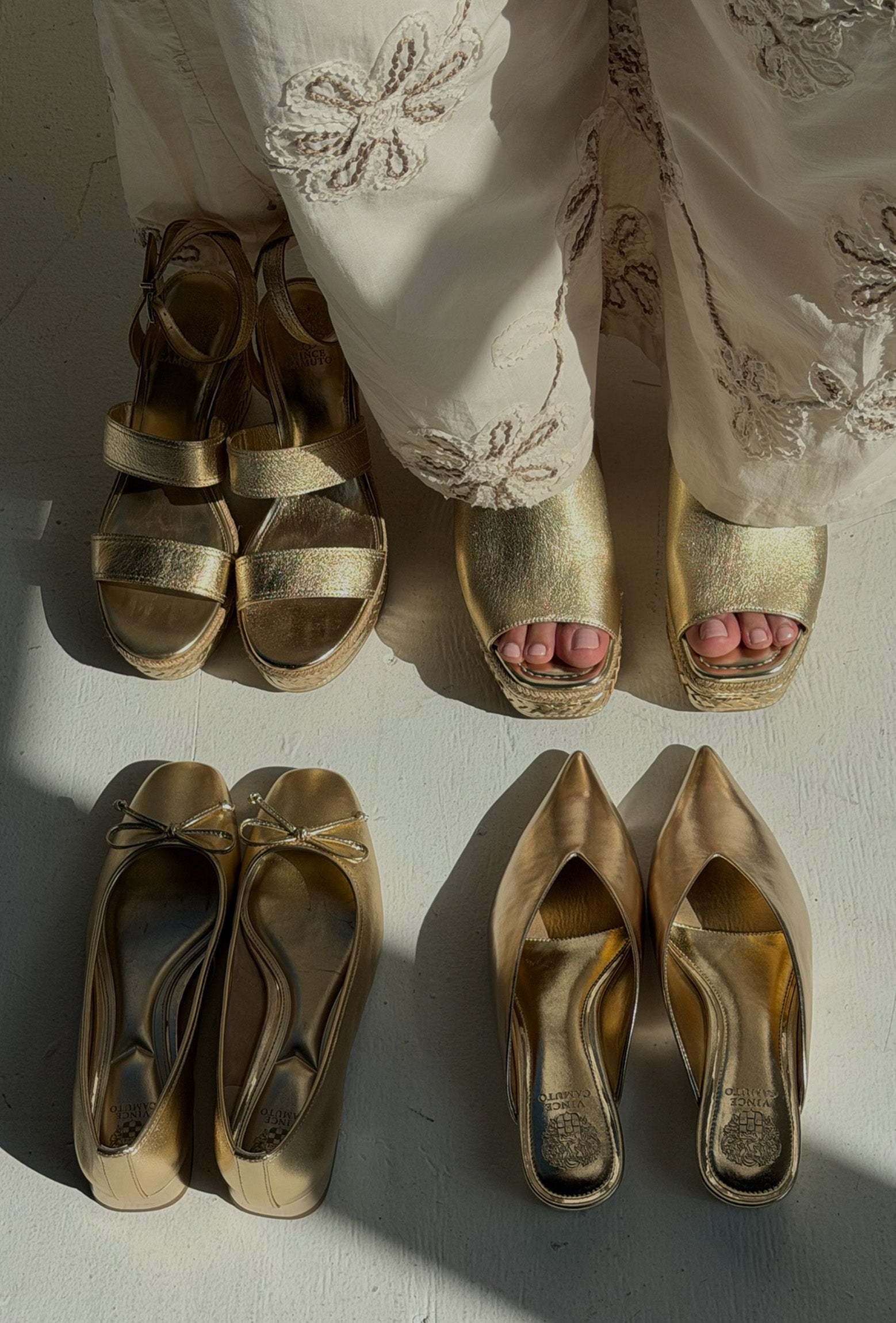 Four pairs of metallic gold women's shoes arranged on a light surface: two platform sandals worn at the top, two pointed flats at the lower left, and two pointed heels at the lower right. A person's lower legs and feet are visible wearing the top pair, so this is classified as Lifestyle.