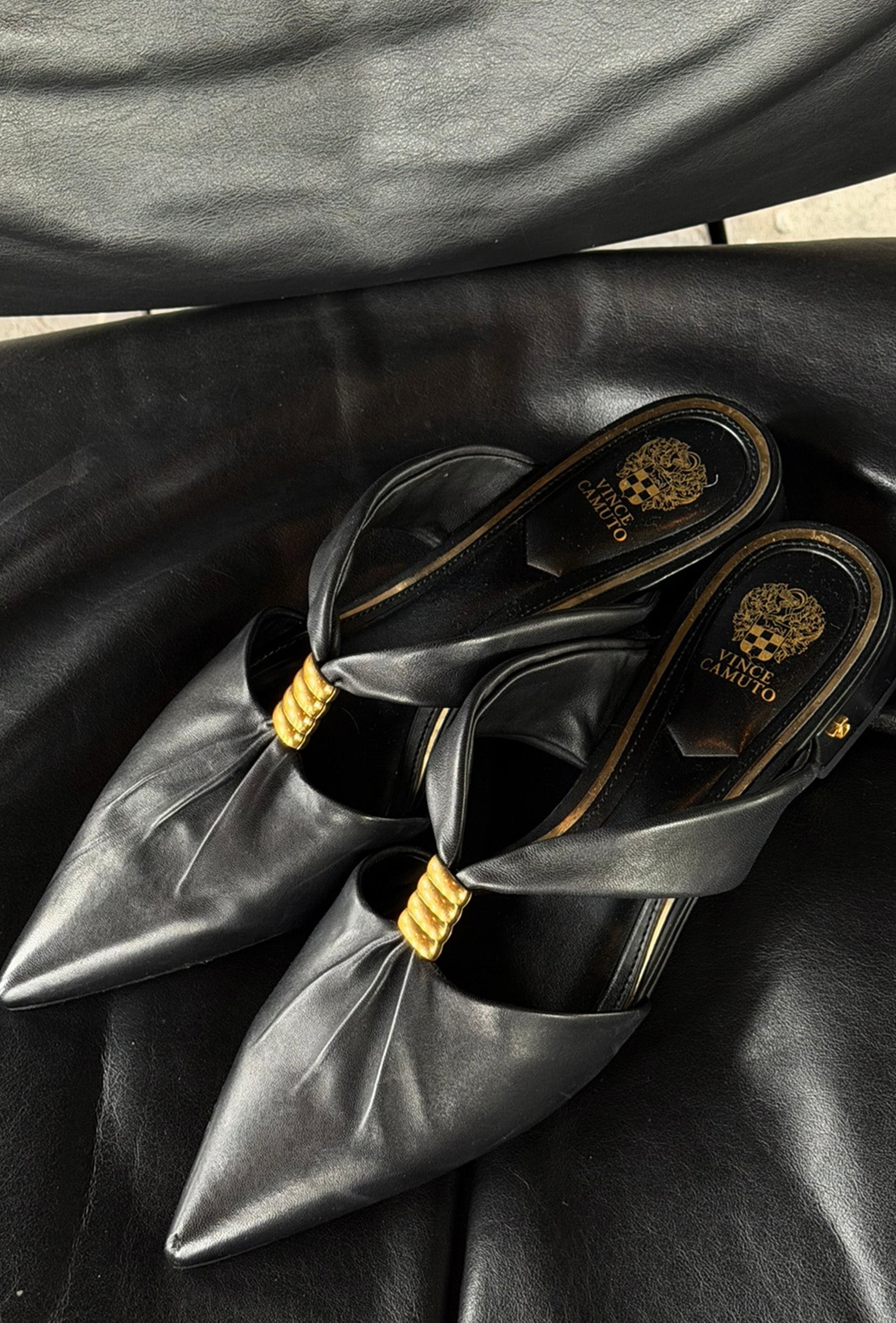 A pair of black leather pointed-toe mule heels with gold hardware and a gold Versace logo on the insole, displayed on a black leather surface.