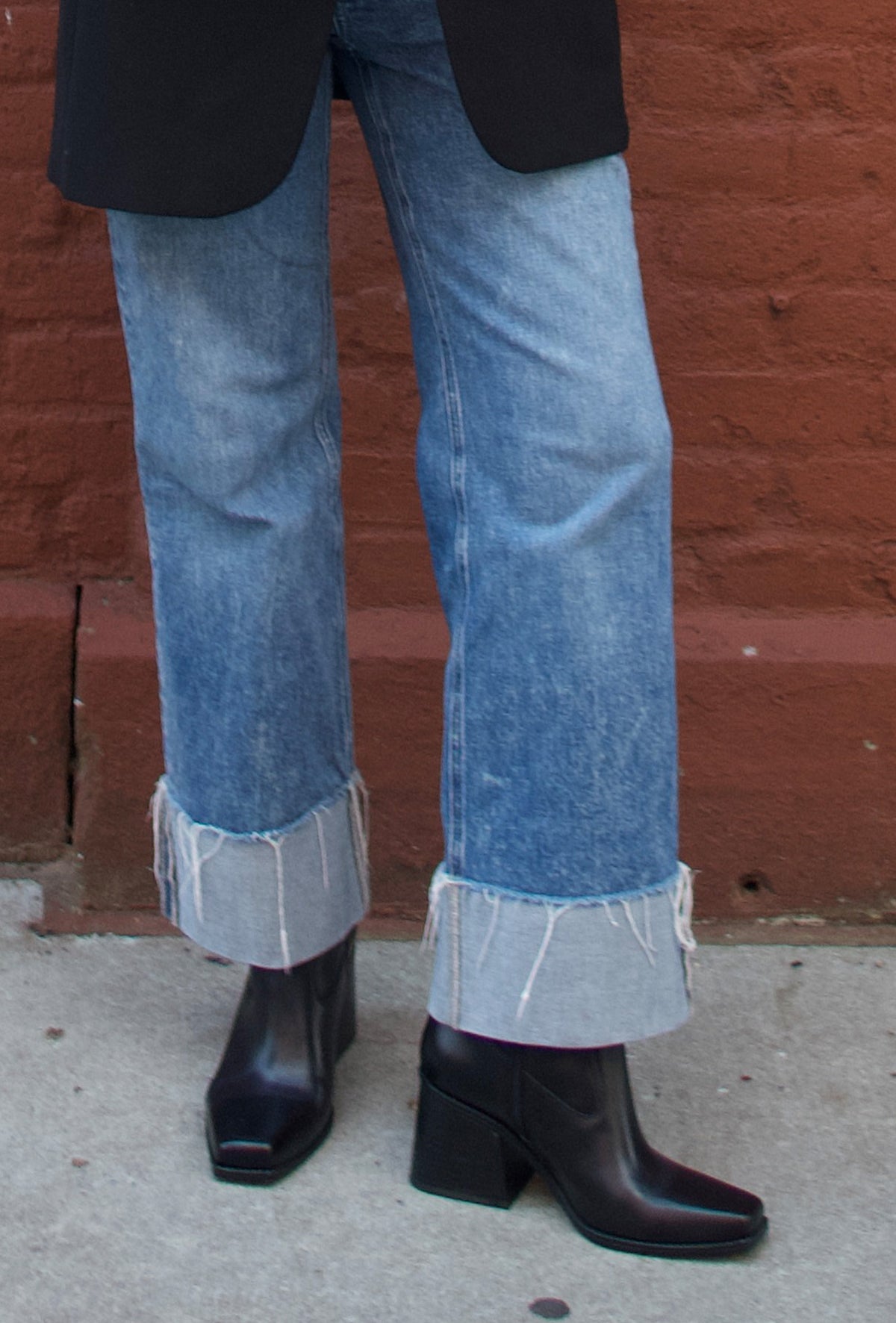 close up of black square toe boots and jeans on a street against a red brick wall 