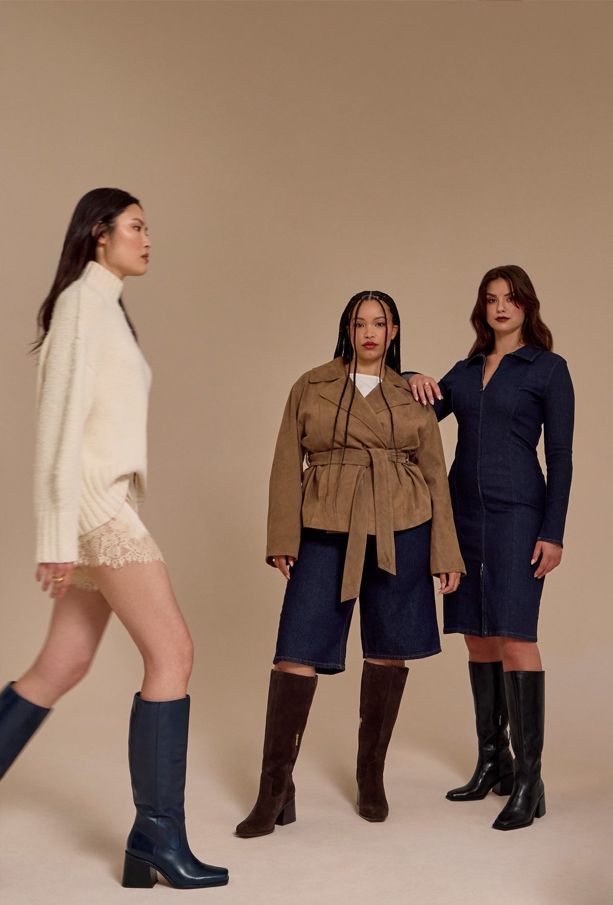 Three women modeling different outfits in a studio setting with a solid beige background. The woman on the left wears a cream knit sweater, lace hem, and dark blue knee-high boots.