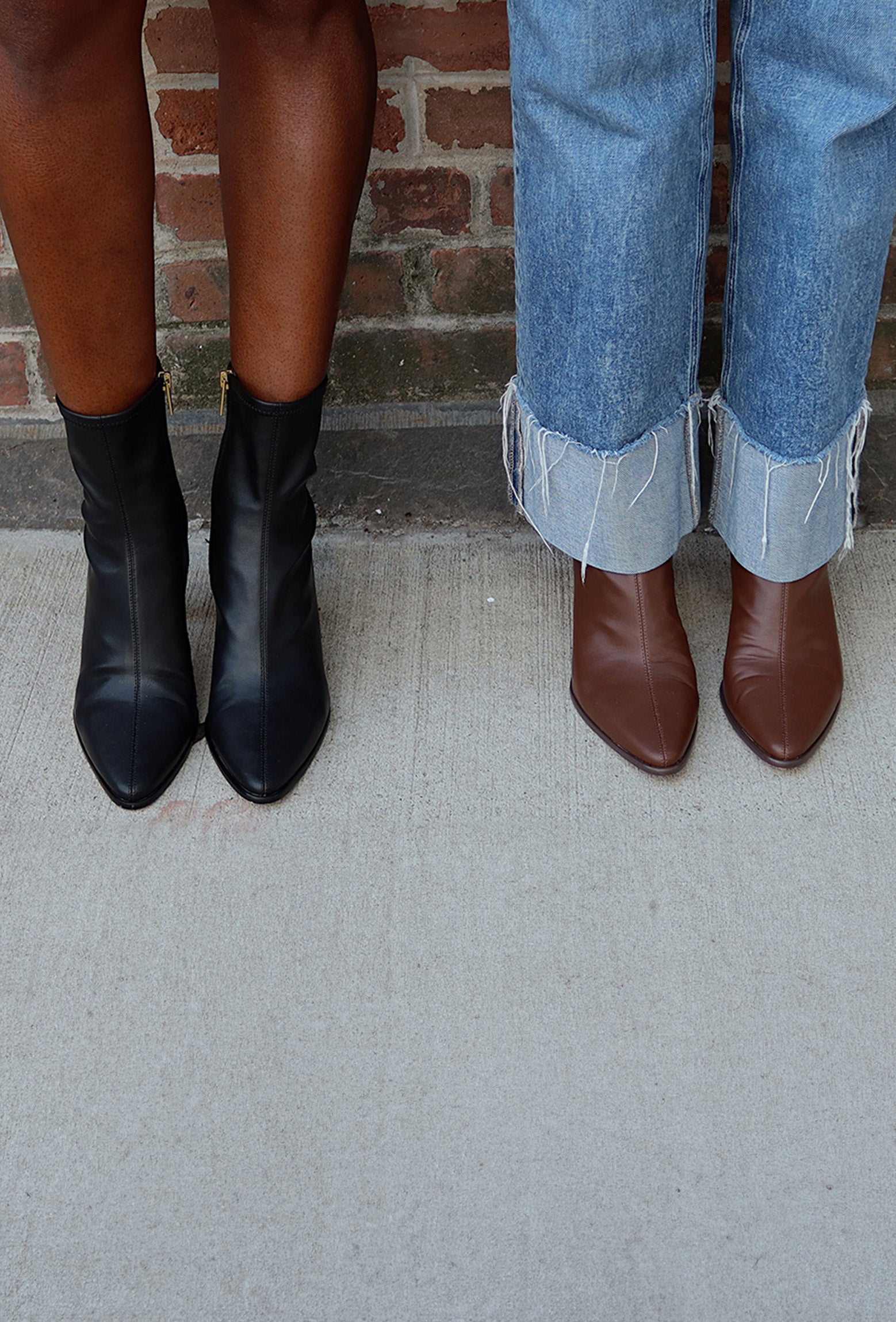 Two people standing side by side outdoors, shown from the knees down, wearing pointed-toe ankle boots. The person on the left wears black leather ankle boots with bare skin visible, and the person on the right wears brown leather ankle boots with cuffed blue jeans.