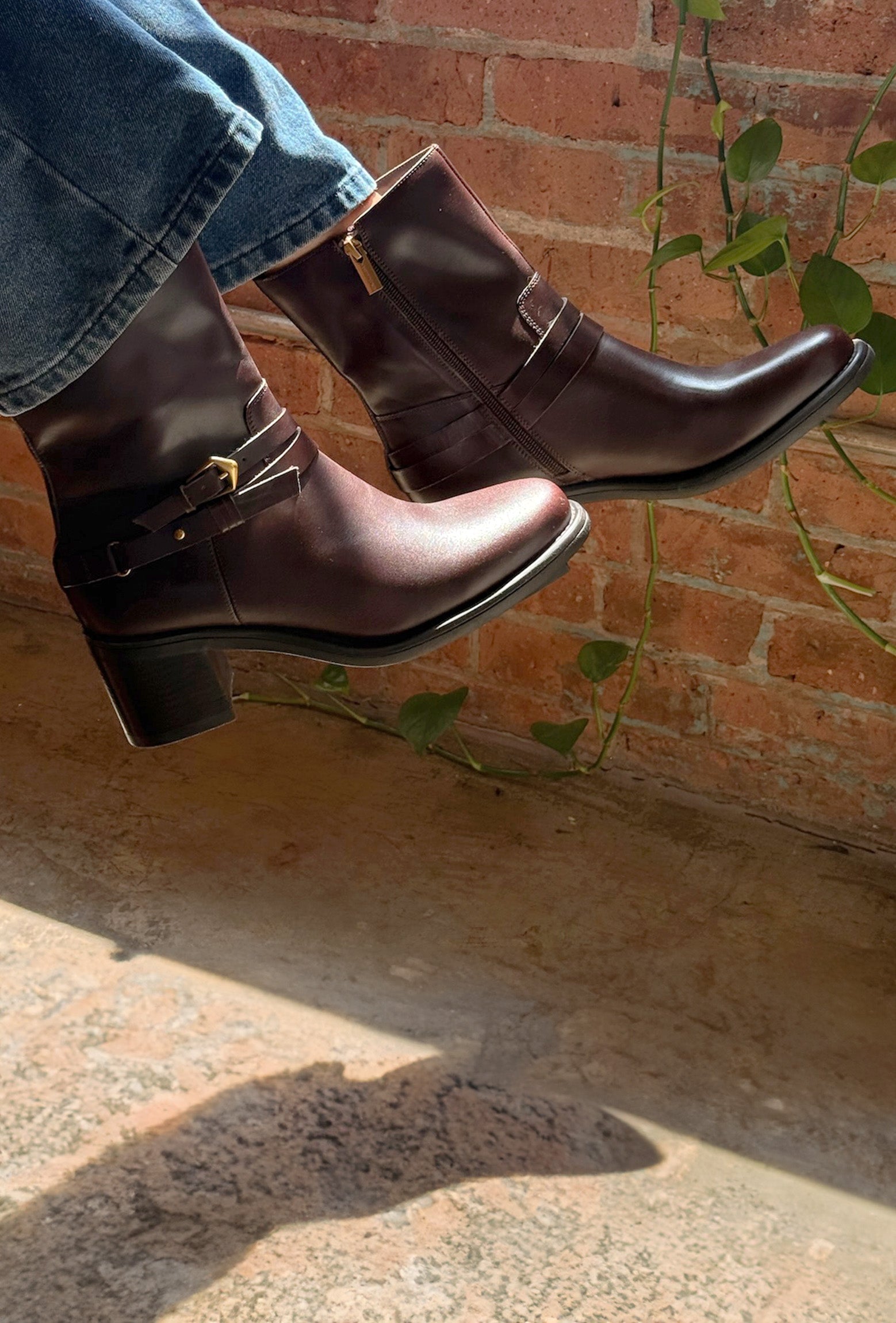 A pair of dark brown leather ankle boots with side buckle straps, worn with blue jeans, shown in a lifestyle setting against a brick wall with climbing green vines.