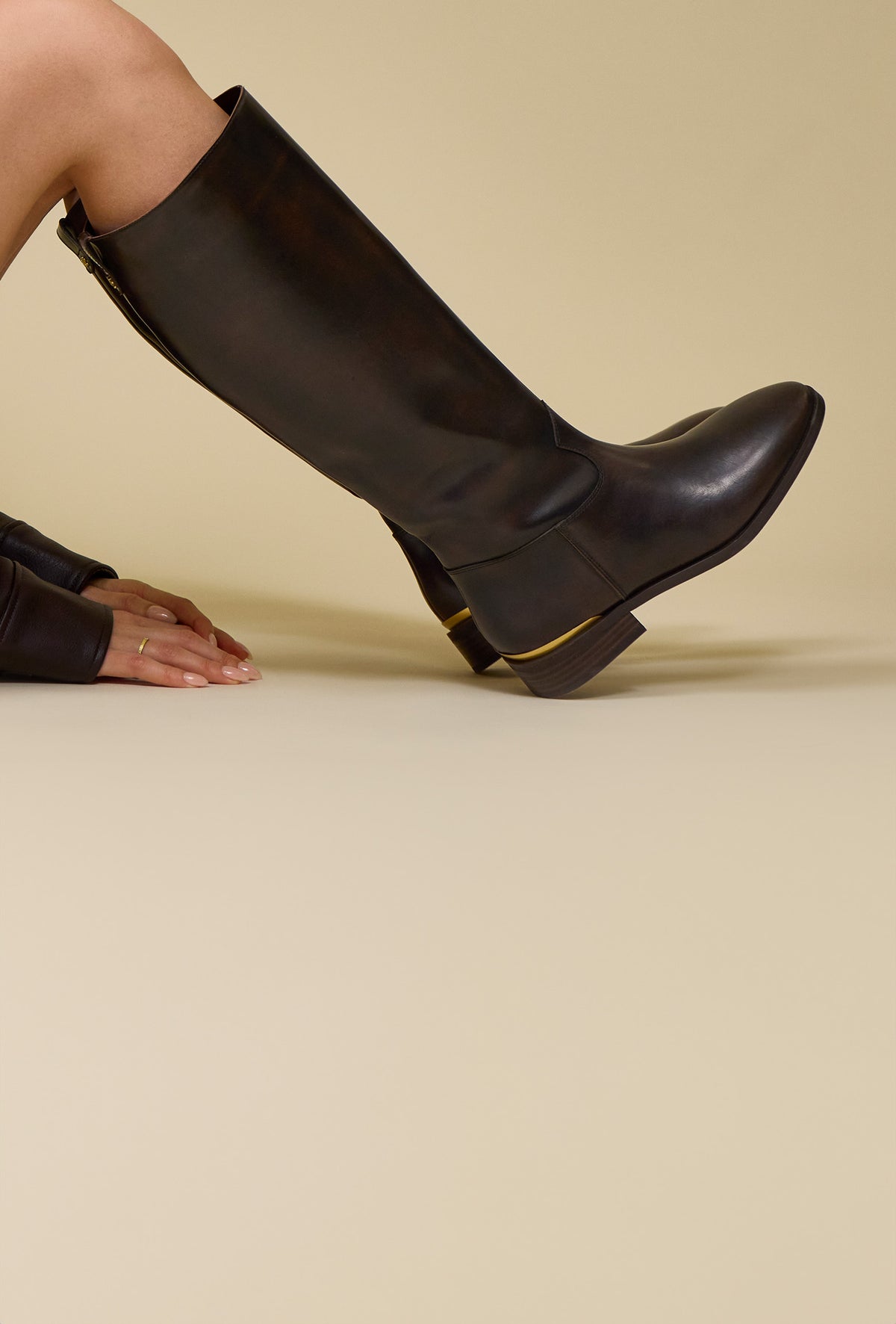 Close-up of woman in dark brown knee-high boots and gold detailing sitting with her knees up against a beige studio background.
