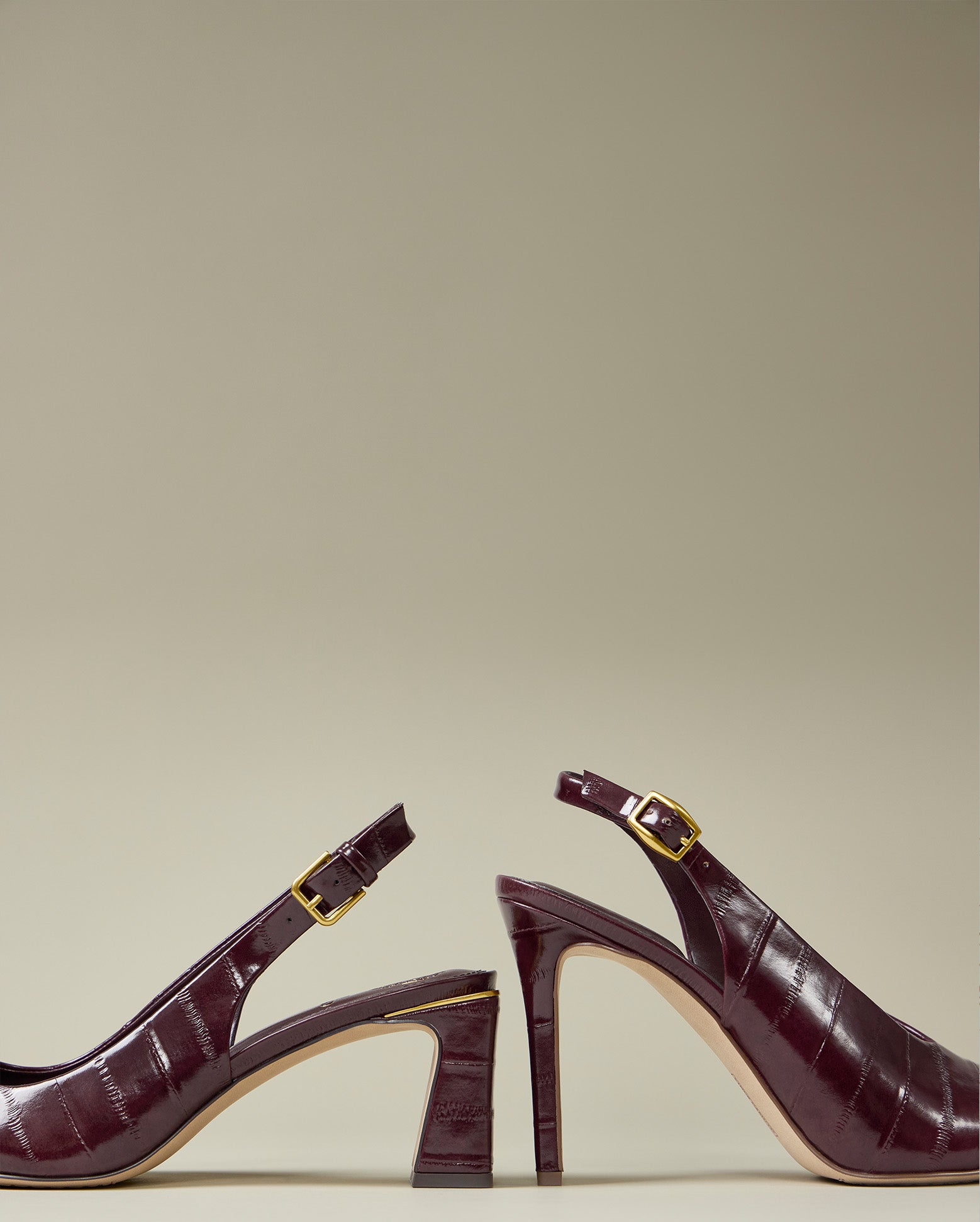 A pair of deep burgundy crocodile-embossed slingback high heels with gold buckle details, shown from side-profile views against a neutral beige background.