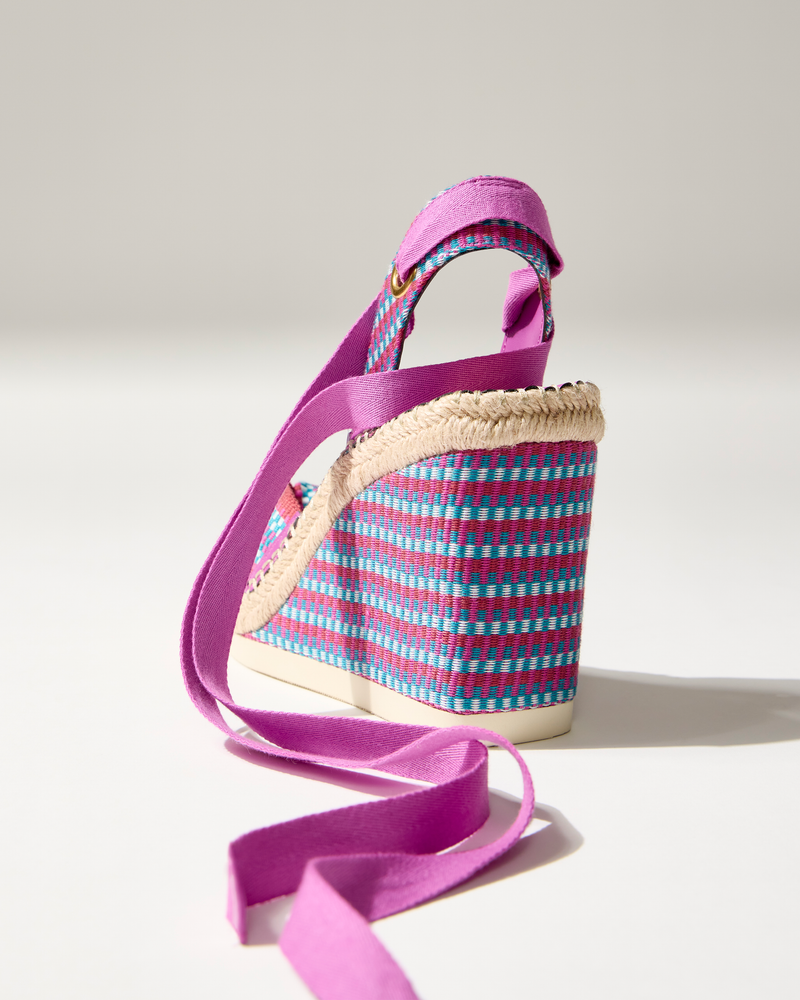 The Bealie Wedge Sandal by Vince Camuto features a bohemian espadrille wedge heel, woven blue and pink pattern, beige rope trim, and long purple ankle tie straps, shown on a white surface in soft lighting.