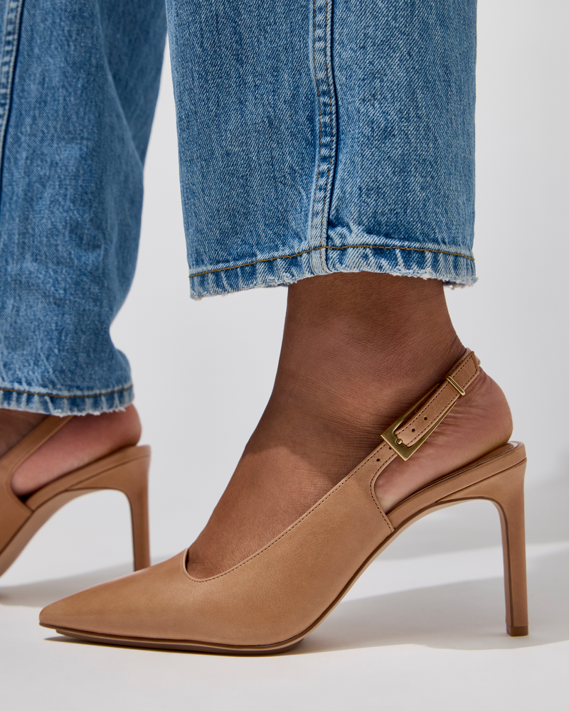 A close-up of a person wearing light blue jeans and Vince Camuto Brendie Pump—beige suede slingback high heel shoes with pointed toes and a gold buckle—standing on a white surface.