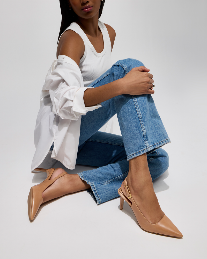 A woman sits on the floor with one knee up, wearing a white tank top, open white shirt, blue jeans, and beige Vince Camuto Brendie Pumps against a plain light background.