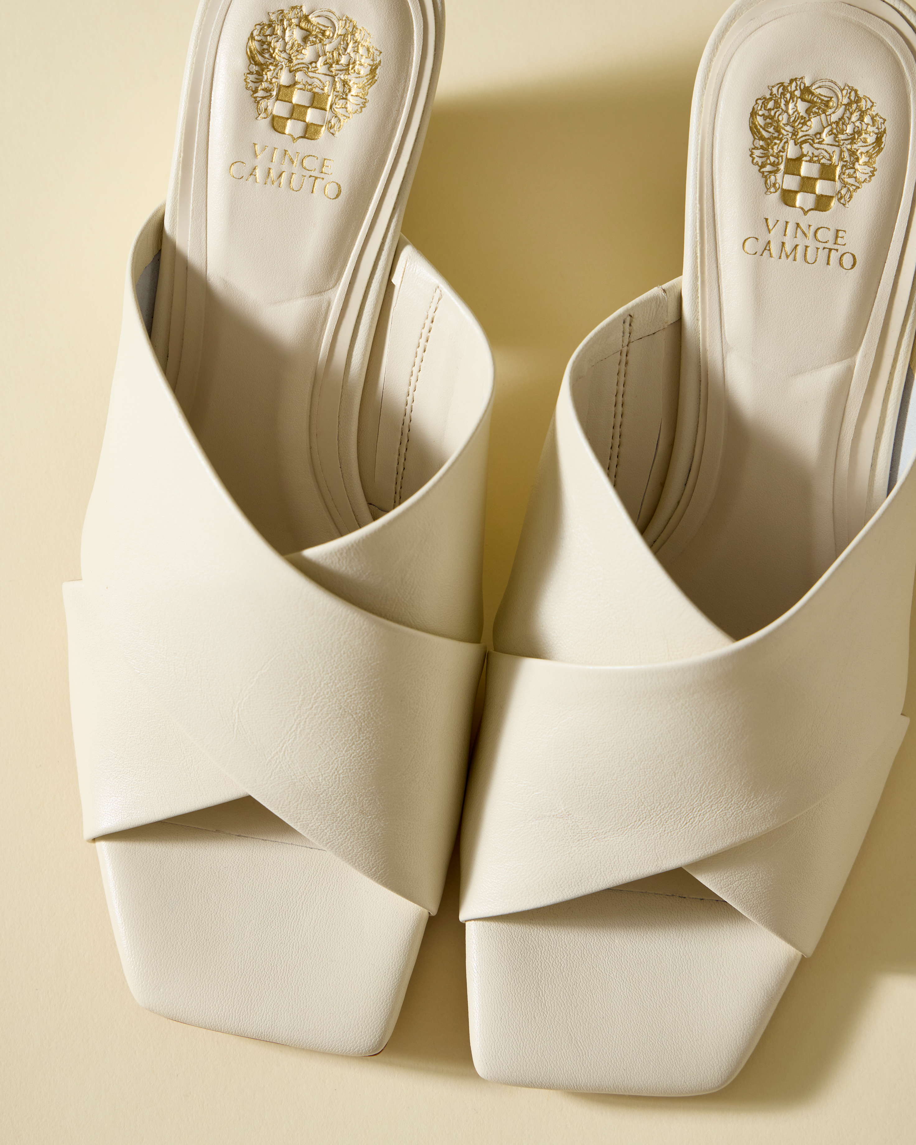 A pair of white Vince Camuto Kespa Mule sandals with a crisscross leather upper and square open toes, displayed side by side on a cream-colored surface.