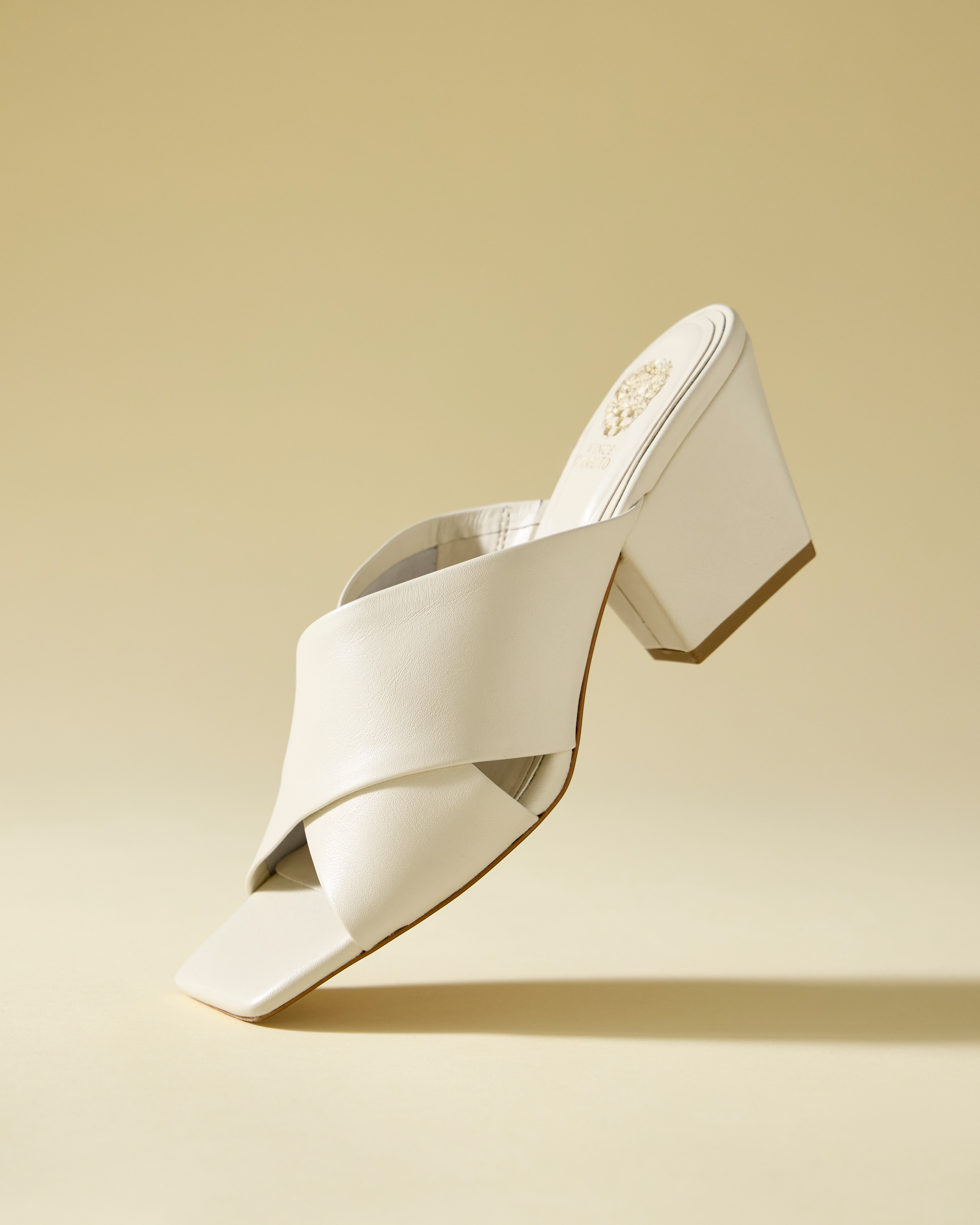 The Kespa Mule by Vince Camuto, a cream-colored block heel sandal with a crisscross leather upper, is displayed floating against a light beige background.
