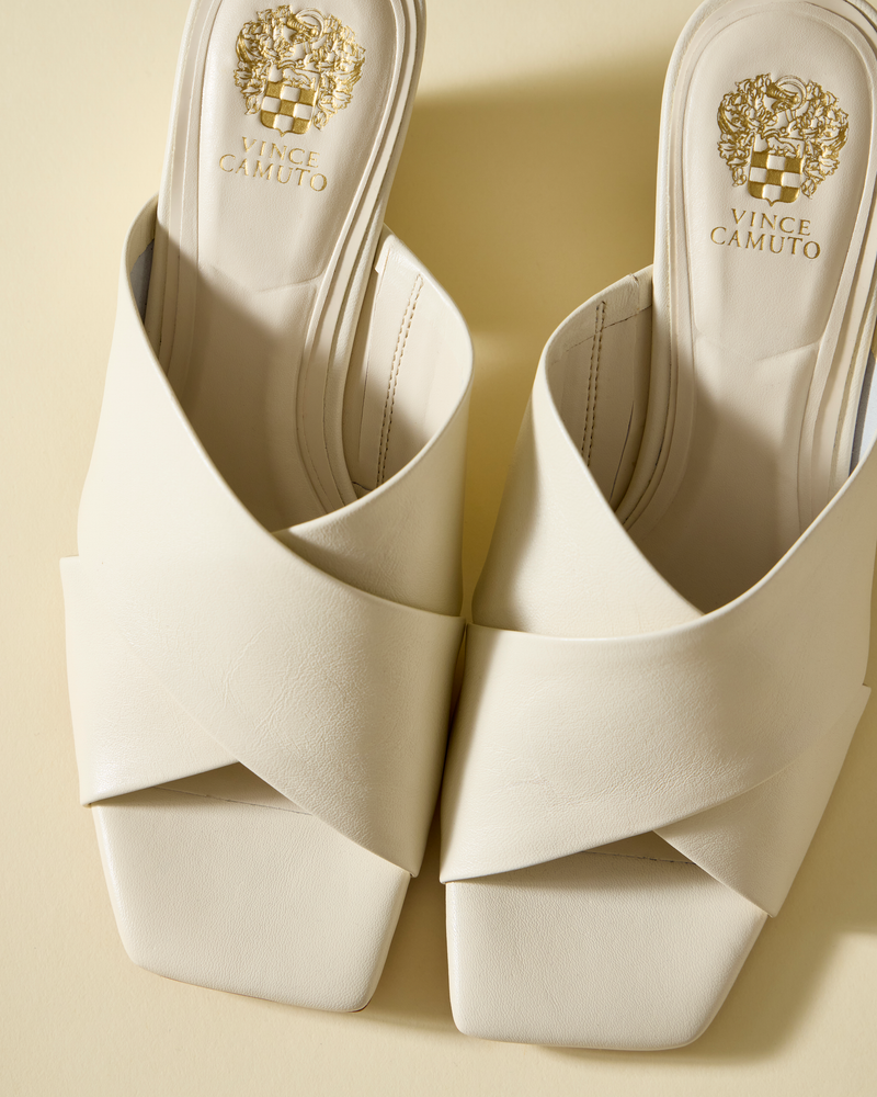 The Vince Camuto Kespa Mule features white leather, crisscross upper, open toes, and square toes. The insole shows the gold Vince Camuto logo. The sandals are displayed on a light beige surface.