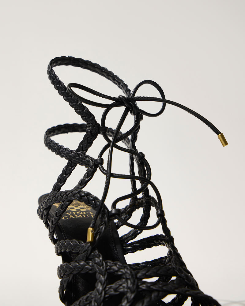 Close-up of the Vince Camuto Tilly Sandal, a black lace-up design with intricate braided straps and gold-tipped laces tied in a bow, set against a plain light background.