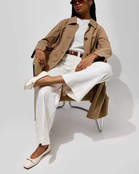 A woman poses confidently on a chair against a plain background, wearing sunglasses, wide-leg pants, a white top, brown belt, tan trench coat with polished hardware, and Vince Camuto's Virna Link Ballet Flat.