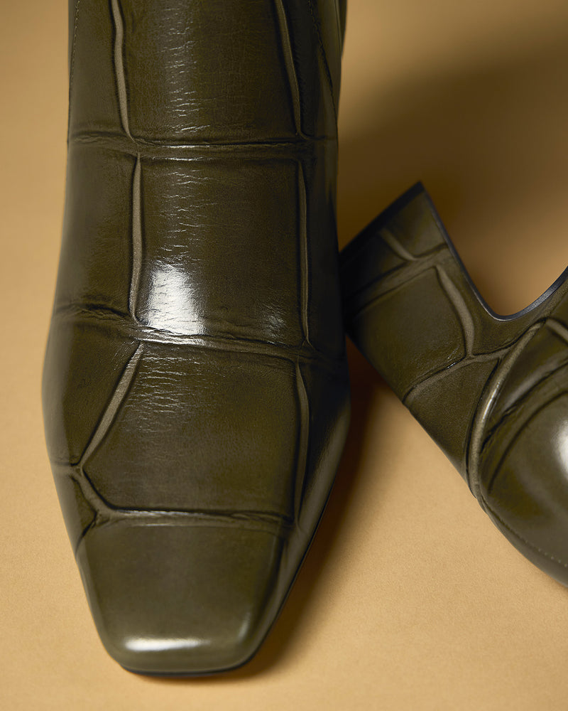 Close-up of two dark green, square-toed Bennidy Knee High Boots by Vince Camuto, featuring crocodile-embossed leather and a stylish block heel, displayed on a tan background.